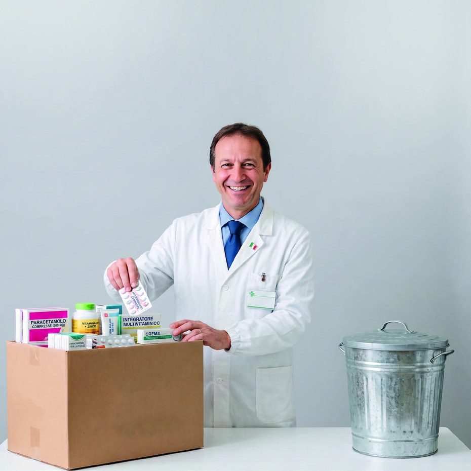 Farmacista in camice bianco che ripone confezioni di farmaci e integratori in uno scatolone, accanto a un bidone: il lavoro sul paniere di smaltimento.